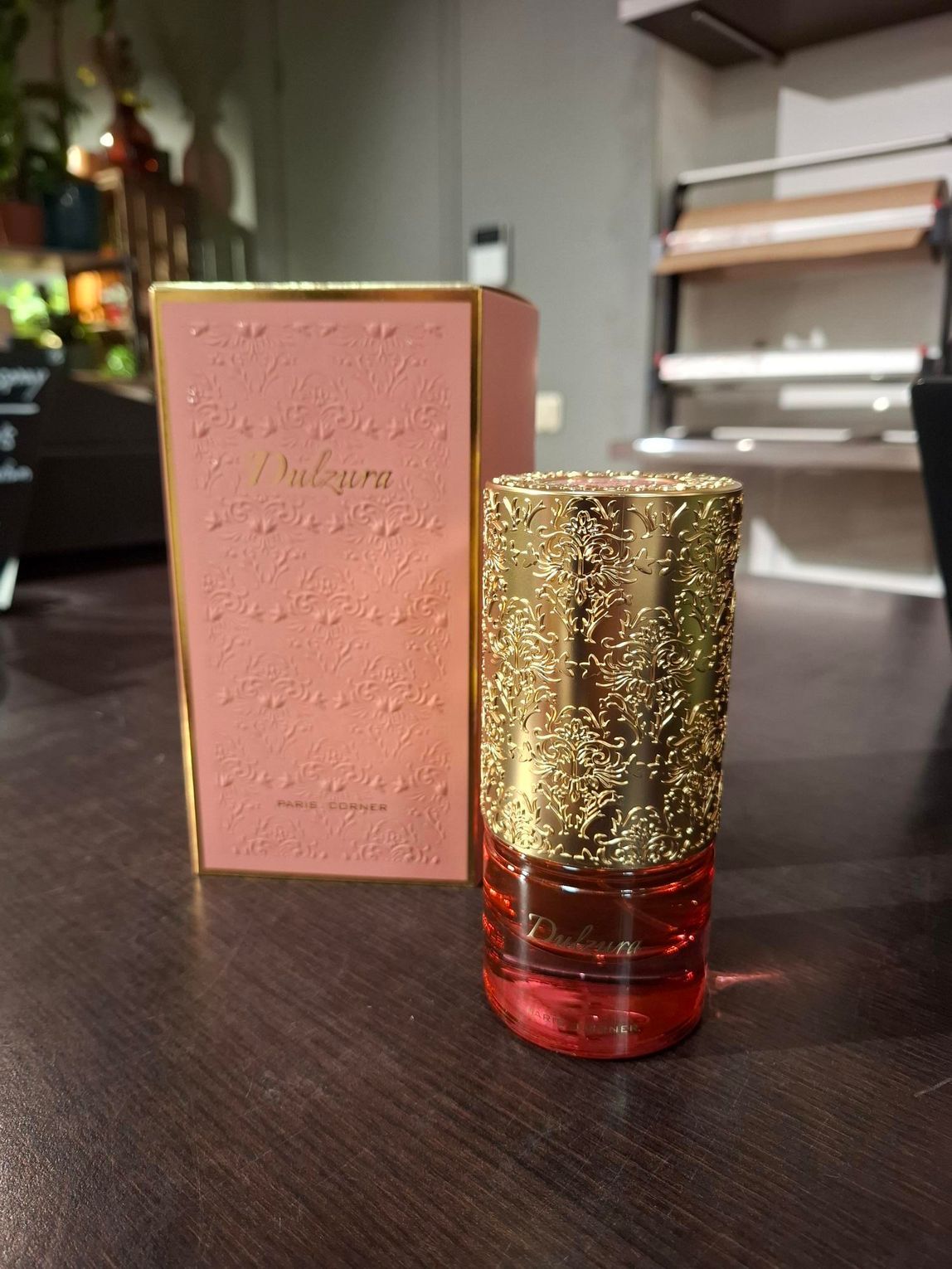 Een roze parfumdoos en een cilindervormige fles met een goudkleurige, gestructureerde dop staan op een donkerhouten ondergrond.