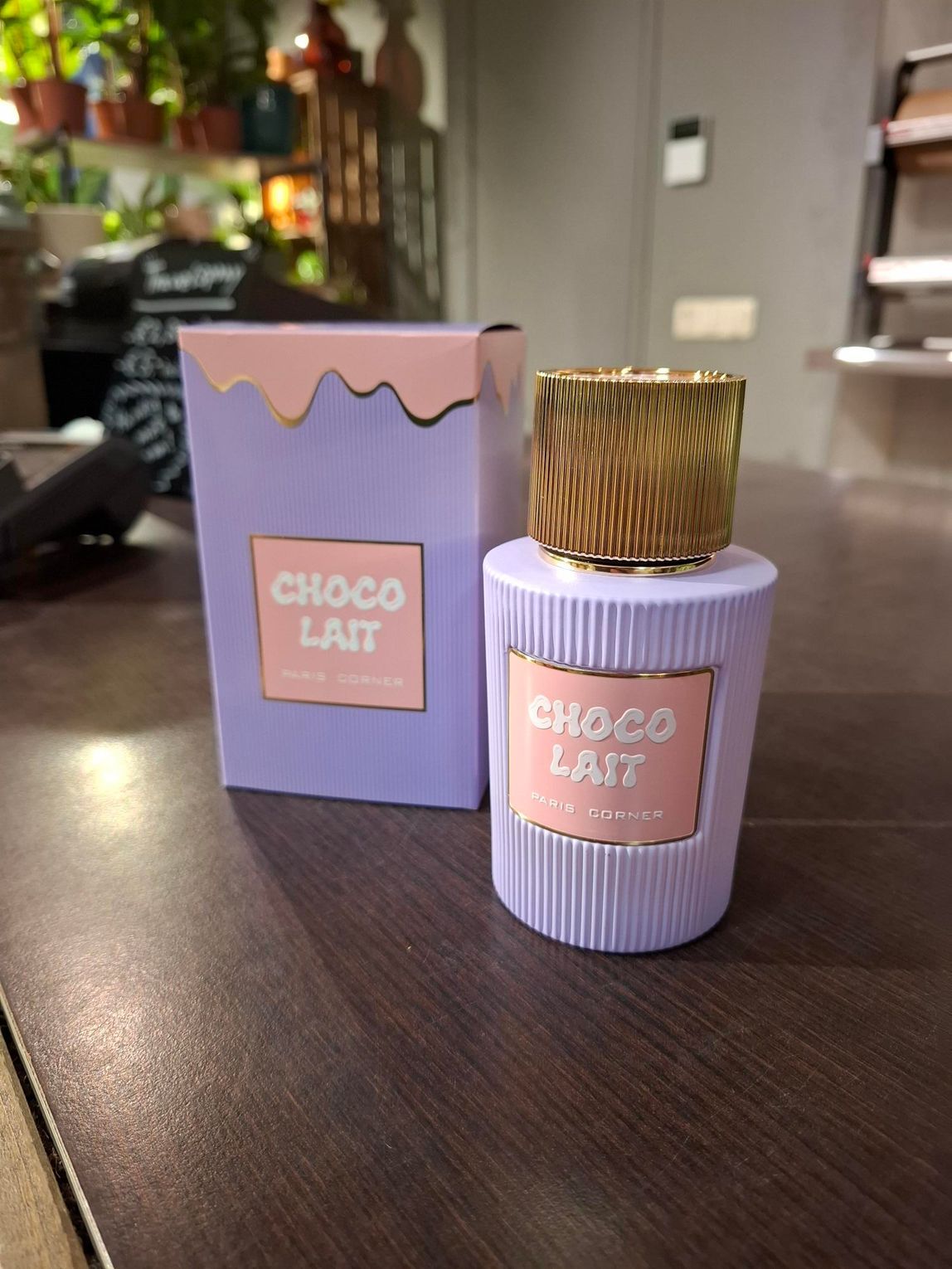 Een flesje Choco Lait-parfum en de bijbehorende doos, beide in pastelpaars met roze etiketten, staan op een houten ondergrond.