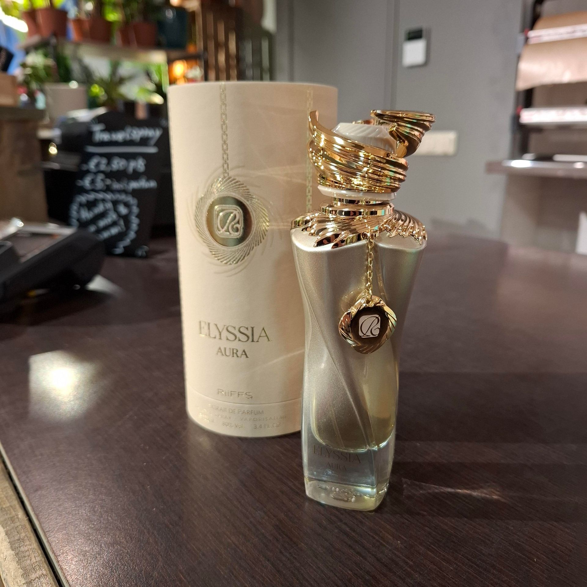 Een flesje Elyssia Aura-parfum en de bijbehorende cilindervormige doos staan op een donkerhouten oppervlak in een zacht verlichte kamer.
