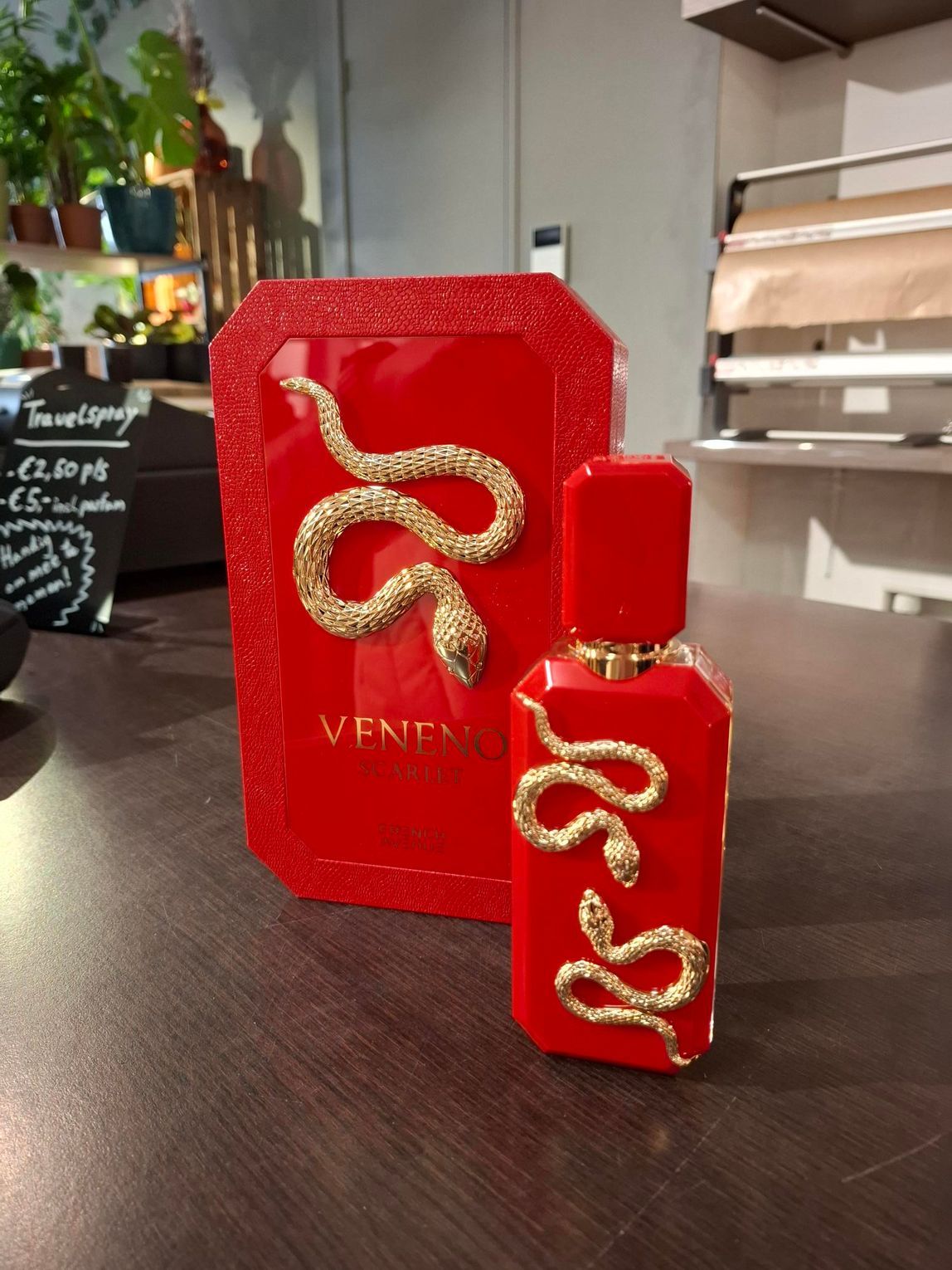 Een rode parfumfles en het bijbehorende doosje, beide versierd met sierlijke gouden slangen, staan uitgestald op een houten ondergrond.