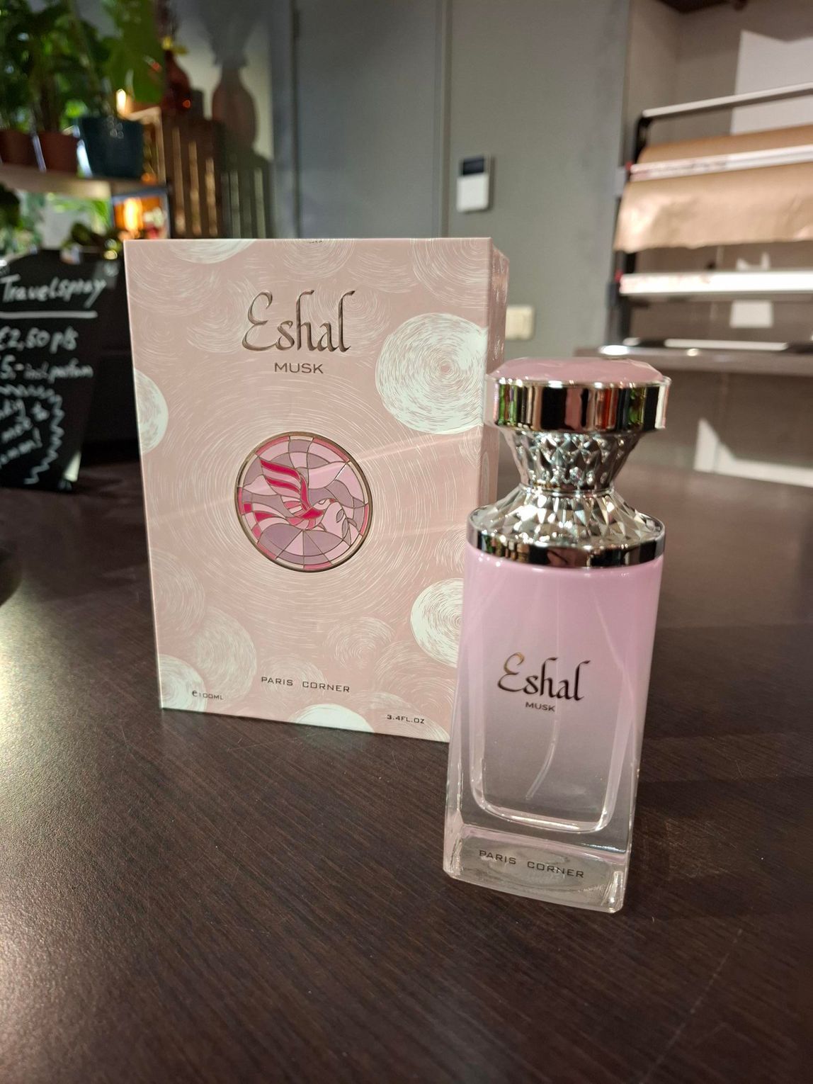 Een flesje roze Eshal-parfum staat voor de bijpassende, geïllustreerde geschenkverpakking met bloemenmotief op een donkere ondergrond.