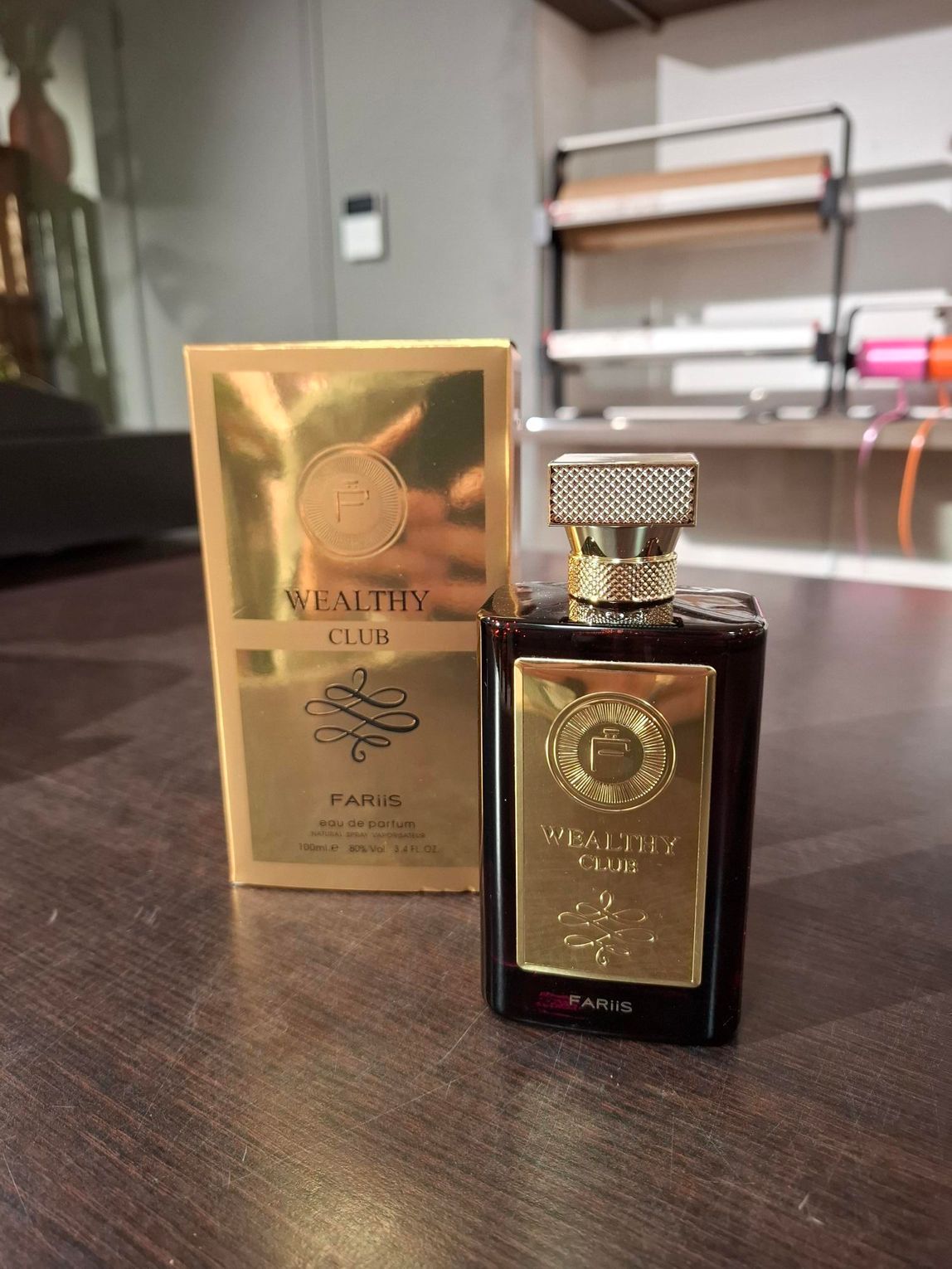 Twee donkere parfumflesjes met gouden etiketten staan op een tafel, waarvan er één in een gouden doosje erachter staat.