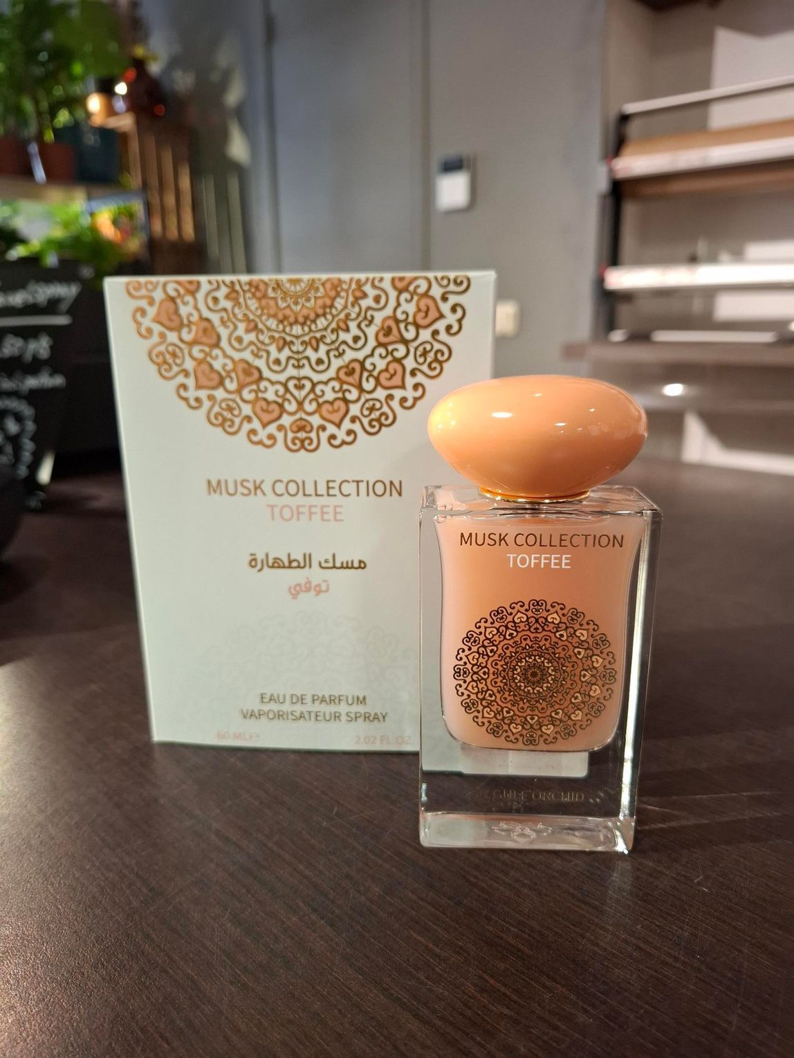Een flesje parfum Musk Collection Toffee en de bijbehorende decoratieve witte doos staan op een houten tafel in een schemerig verlichte ruimte.