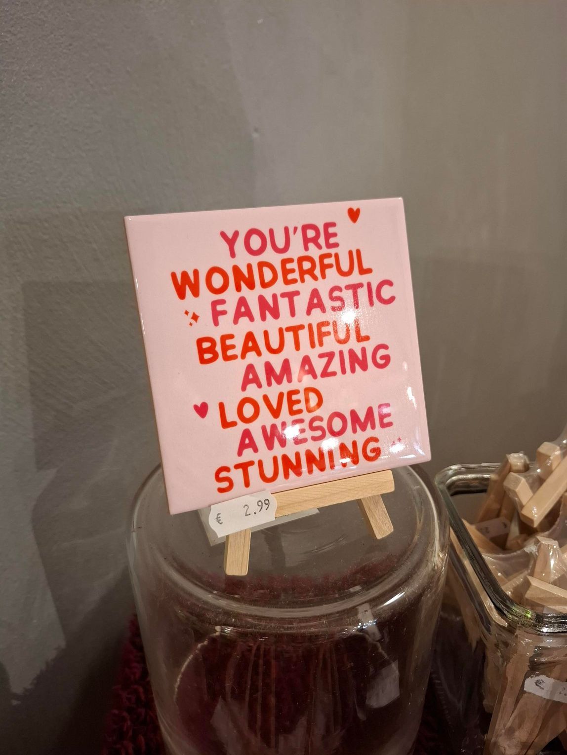 Een hand die een kleine houten katapult vasthoudt in een winkelvitrine met diverse houten speeltjes en een roze bord.