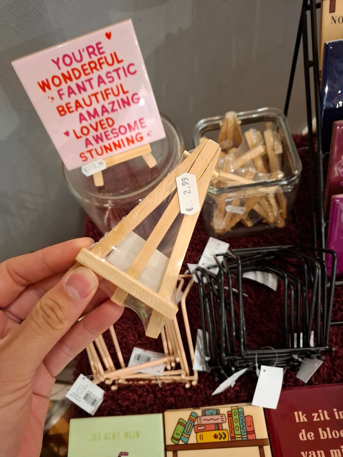 Een hand die een kleine houten katapult vasthoudt in een winkelvitrine met diverse houten speeltjes en een roze bord.