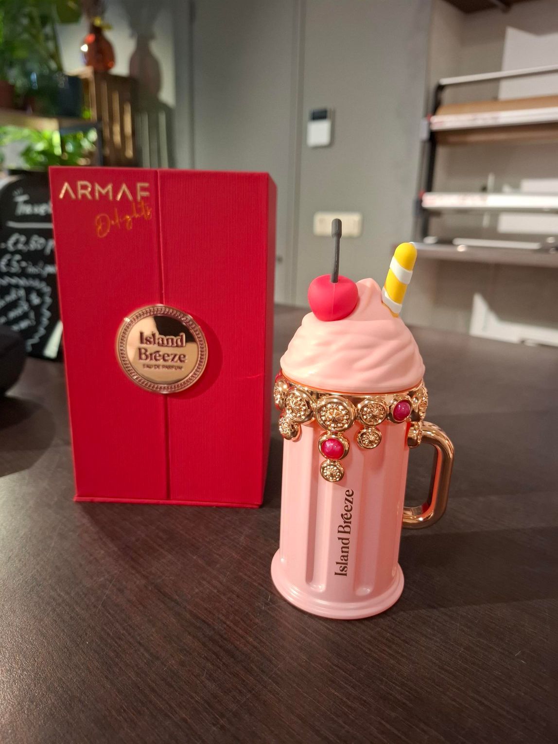 Een roze parfumflesje in de vorm van een milkshake met een kers en een rietje ernaast, naast een rode ARMAF-doos op een tafel.