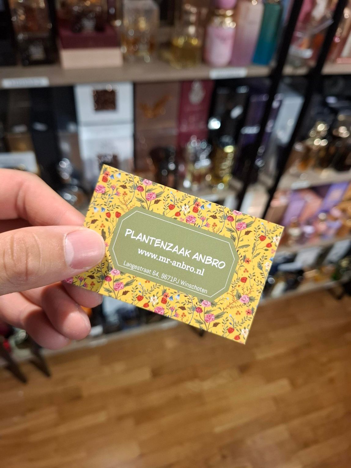 Hand die een kleurrijk visitekaartje vasthoudt in een winkelgang met schappen vol flessen op de achtergrond.
