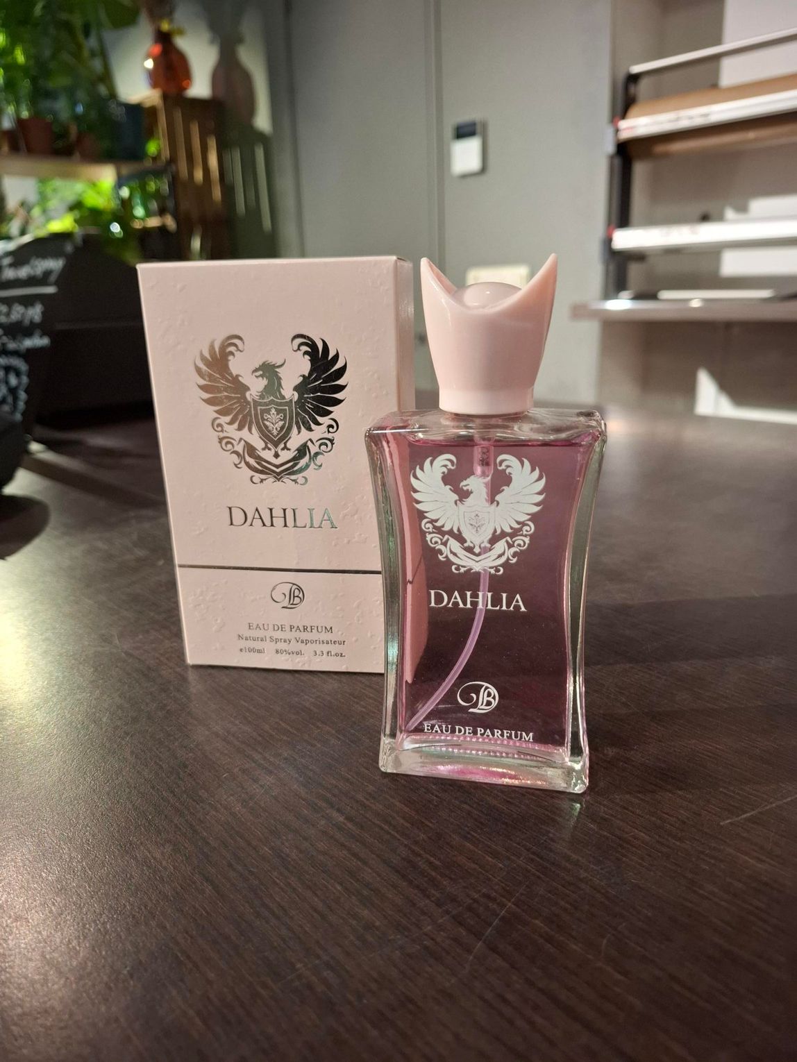 Het flesje en doosje van het parfum Pink Dahlia staan op een donkere tafel in een lichte kamer.