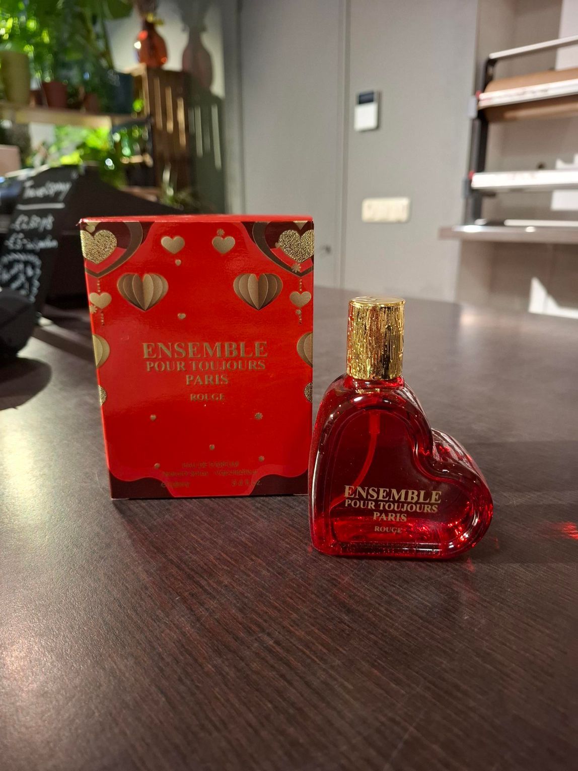 Een rood, hartvormig parfumflesje met gouden dop, naast een bijpassende rode geschenkdoos op een donkere tafel.