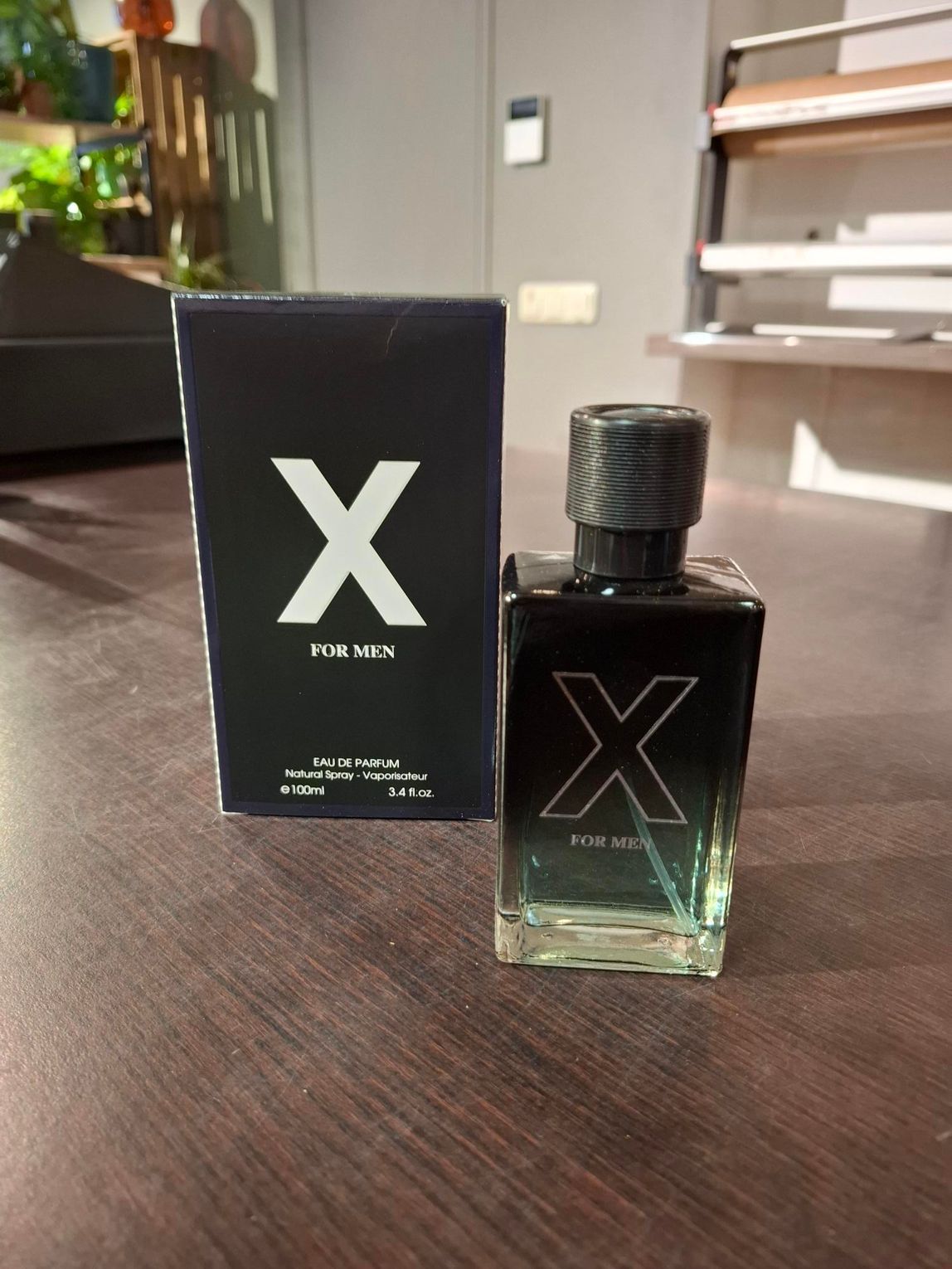 Zwarte parfumfles met een X-logo naast de bijbehorende doos op een houten tafel.