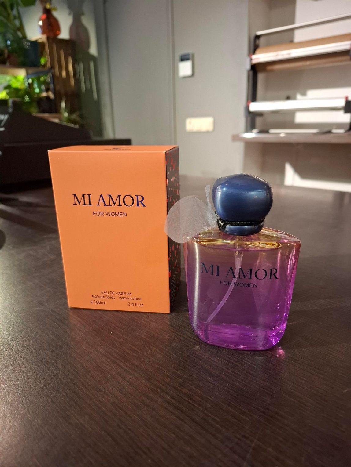 Paarse parfumfles met blauwe dop naast een oranje M'Amor-doos op een tafel.