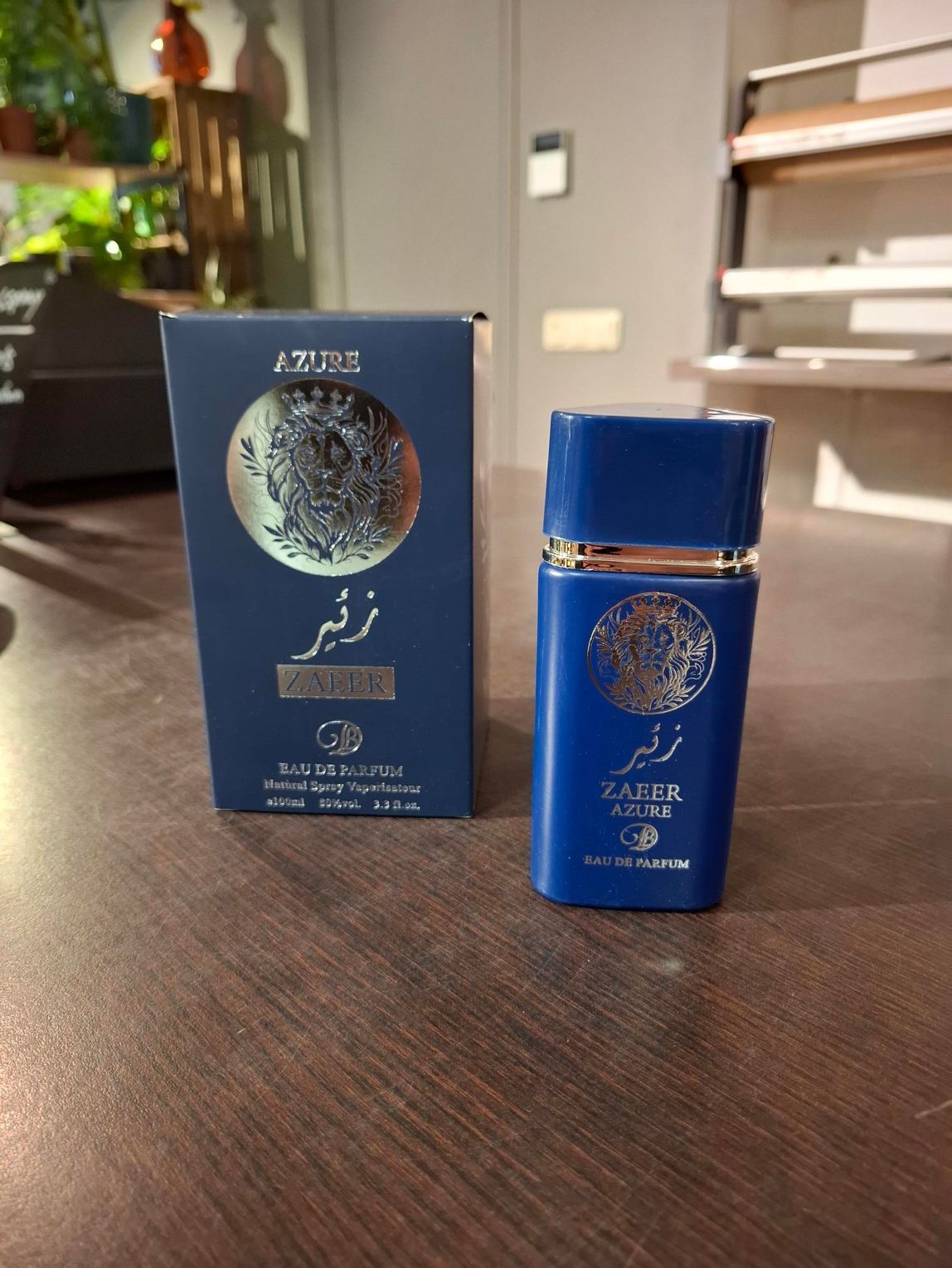 Een blauw parfumflesje met bijpassend doosje op een houten tafel binnenshuis, met een raam op de achtergrond.