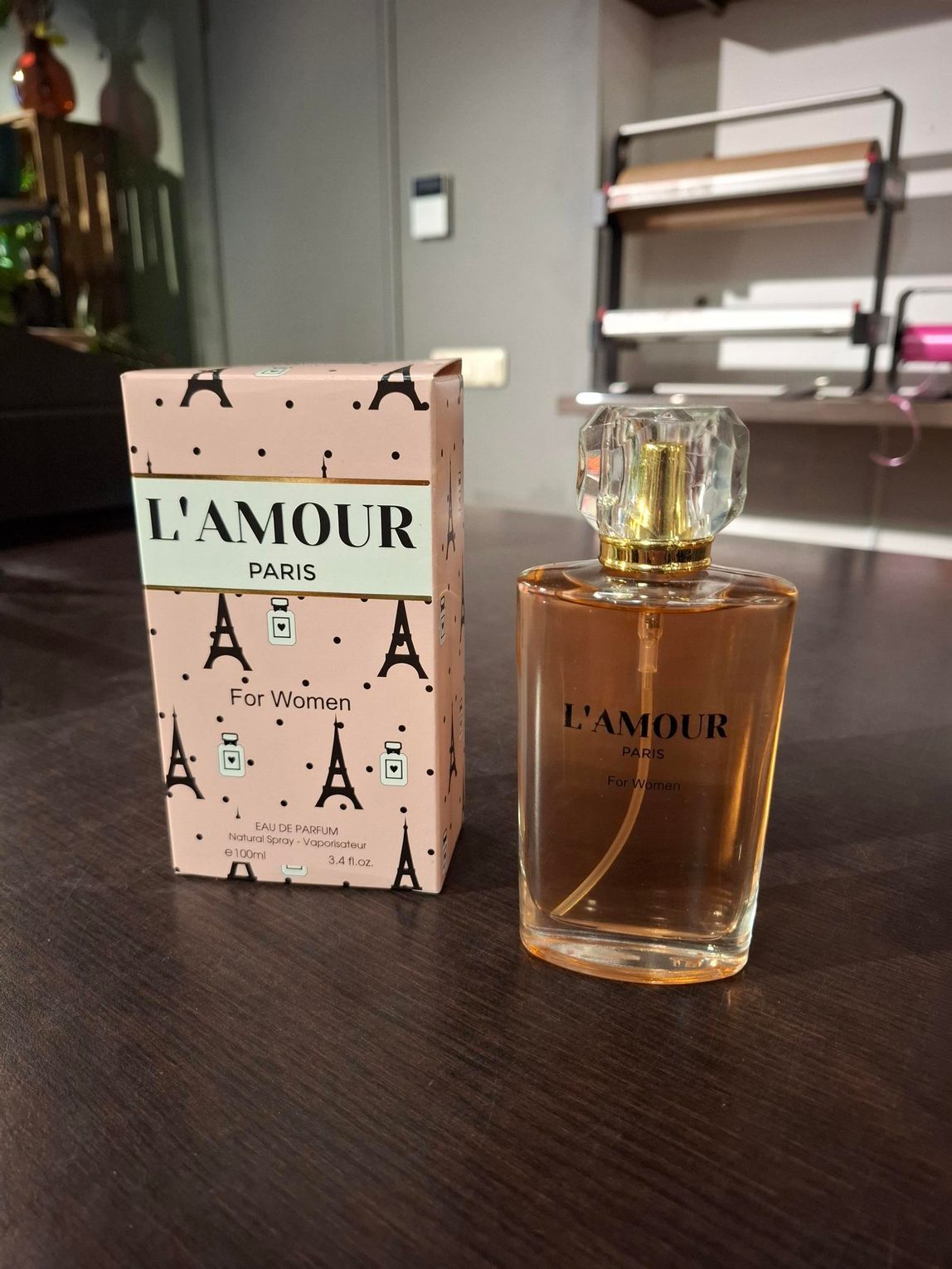 Het flesje en doosje van het parfum L'Amour staan op een donkere tafel in een zacht verlichte kamer.