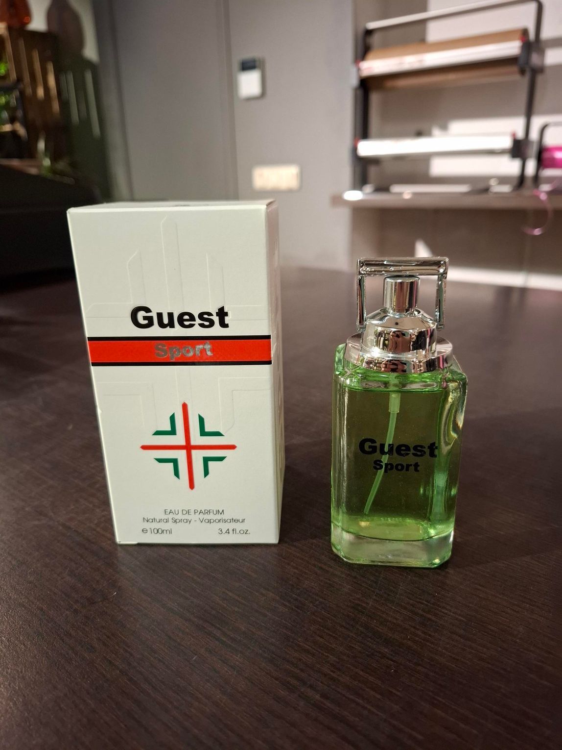 Een flesje parfum van Guest Staff, met groene vloeistof erin, staat naast de witte doos op een tafel binnen.