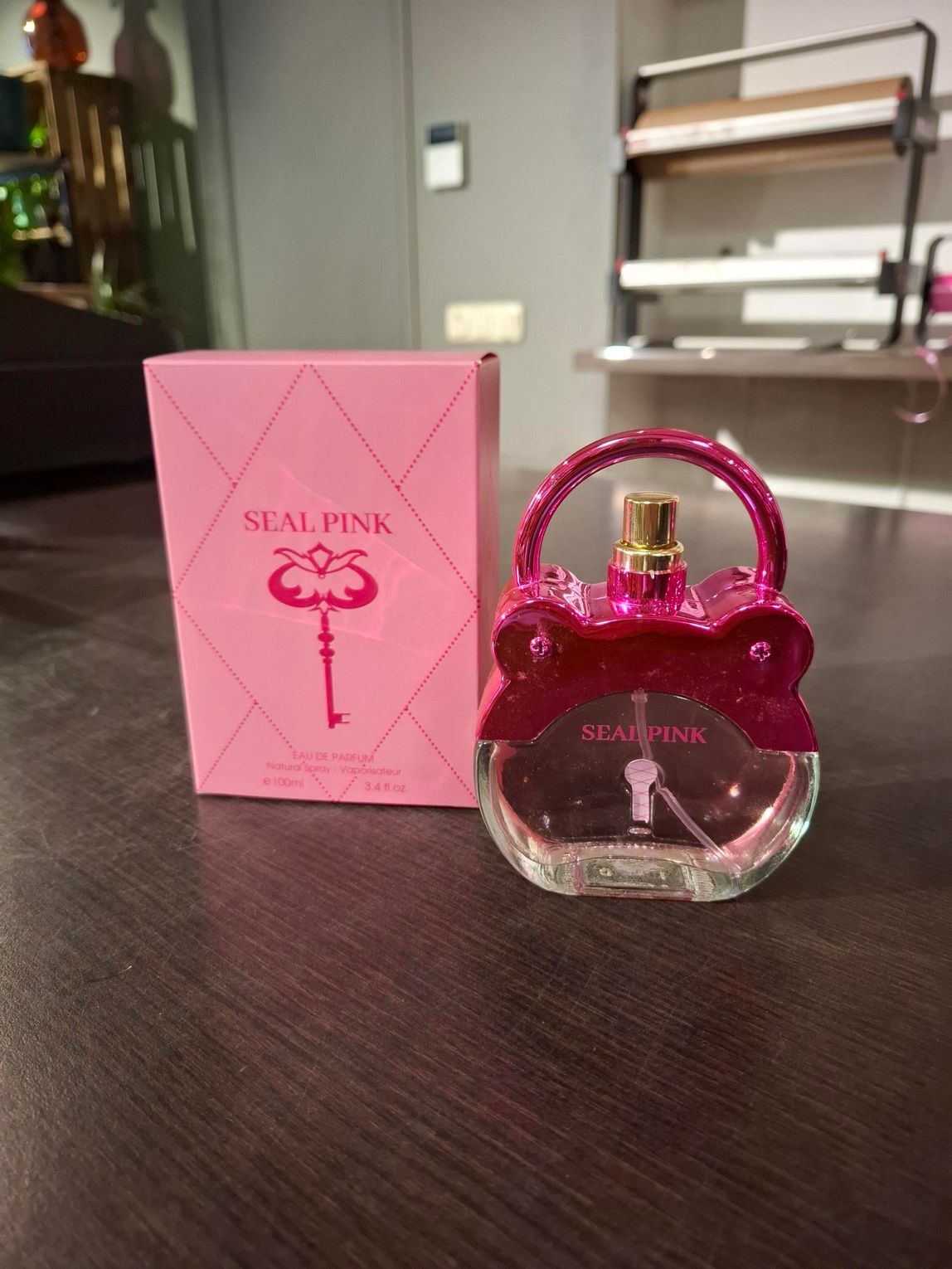 Een roze parfumflesje met bijpassend doosje op een donkere tafel, met een wazige kamer op de achtergrond.