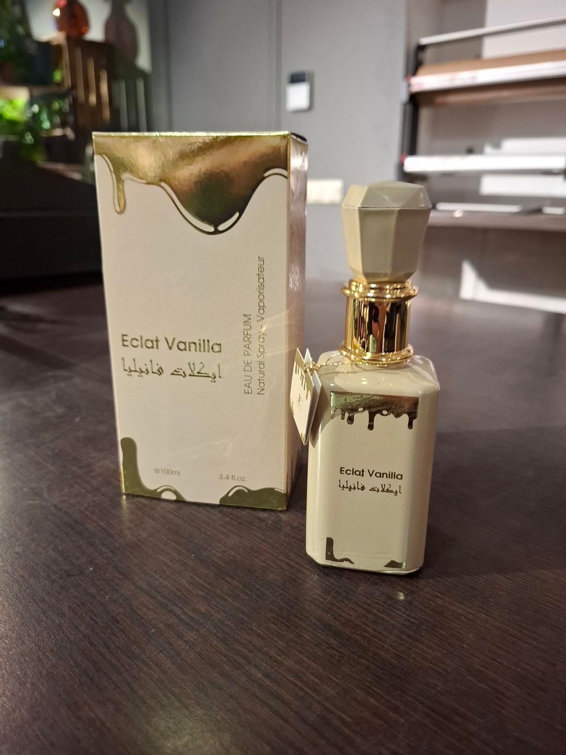 Parfumflesje met gouden dop naast de bijpassende crèmekleurige en gouden doos op een donkere tafel.