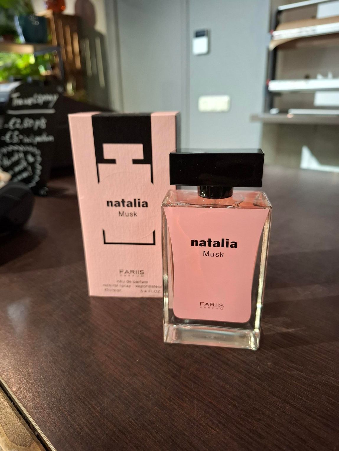 Roze Natalia parfumflesje en -doos op een houten tafel, met een wazige caféachtergrond.