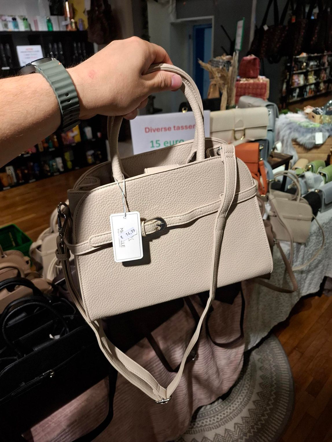 Beige handtas met schouderband, tentoongesteld in een winkeldisplay, met een prijskaartje eraan.