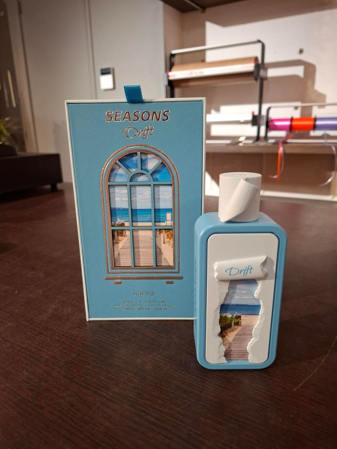 Een blauwe parfumfles met bijpassend doosje op een tafel, met op de achtergrond een wazig beeld van een kamerinterieur.