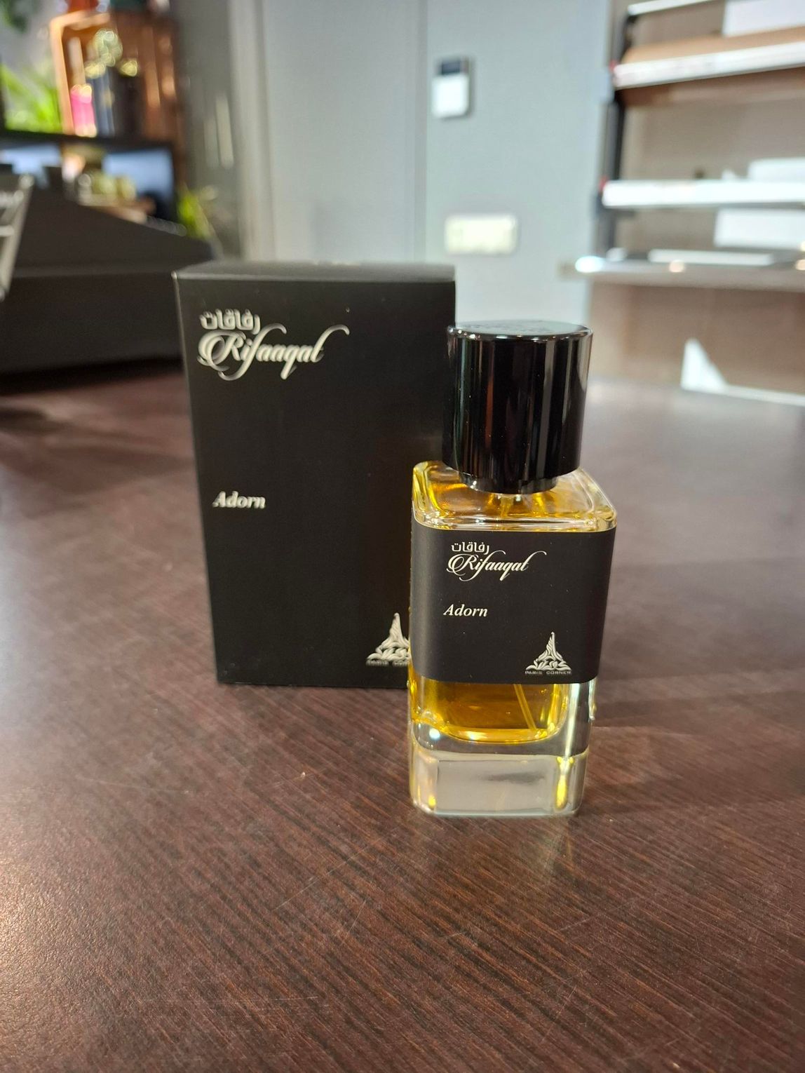 Klein amberkleurig parfumflesje met zwarte dop, naast een bijpassende zwarte doos op een houten tafel.