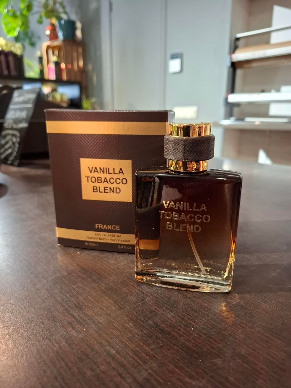 Parfumflesje en -doos van Vanilla Tobacco Blend op een houten tafel, met een wazige caféachtergrond.