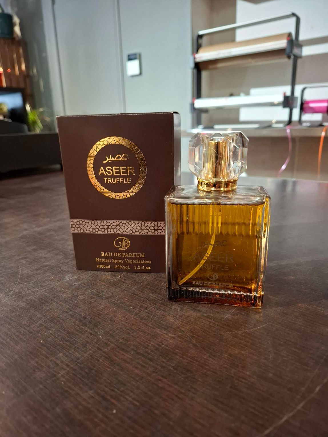 Een flesje amberkleurige parfum naast de bruine doos op een donkere tafel in een moderne kamer.