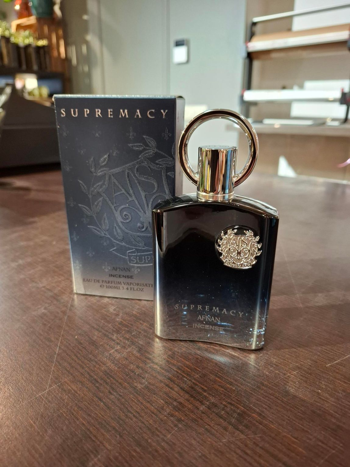 Zwarte parfumfles met gouden dop naast een Supremacy-doos op een houten vloer.