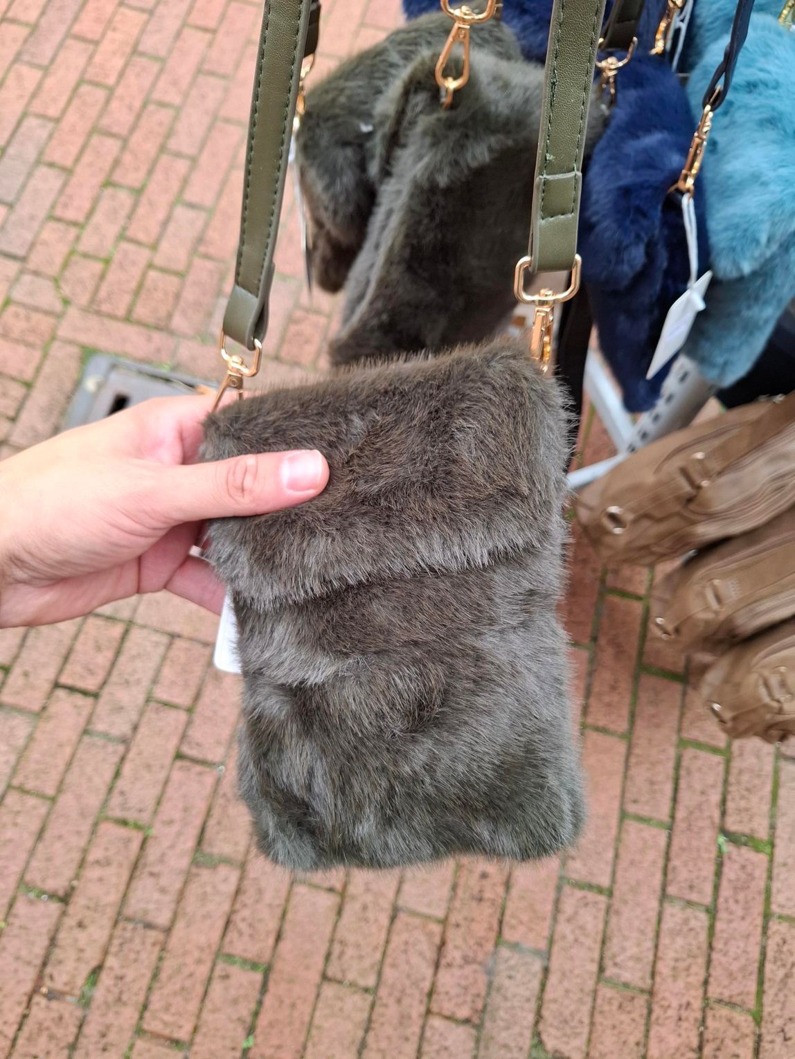 Hand die een grijze bonten tas met een schouderband vasthoudt op een stenen pad.