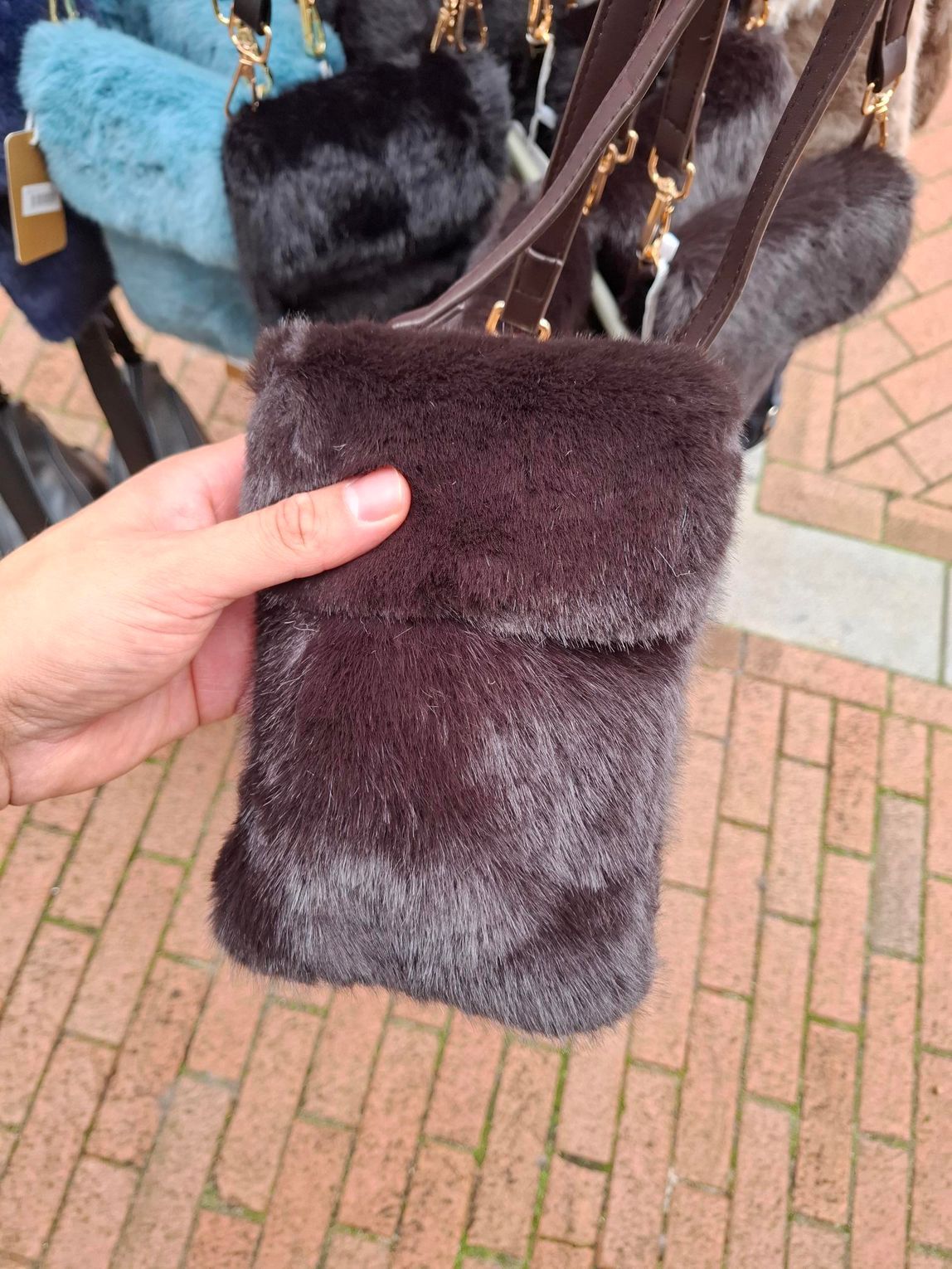 Hand met een klein donkerbruin tasje van imitatiebont met een gouden kettingriem, buiten op een bakstenen stoep.
