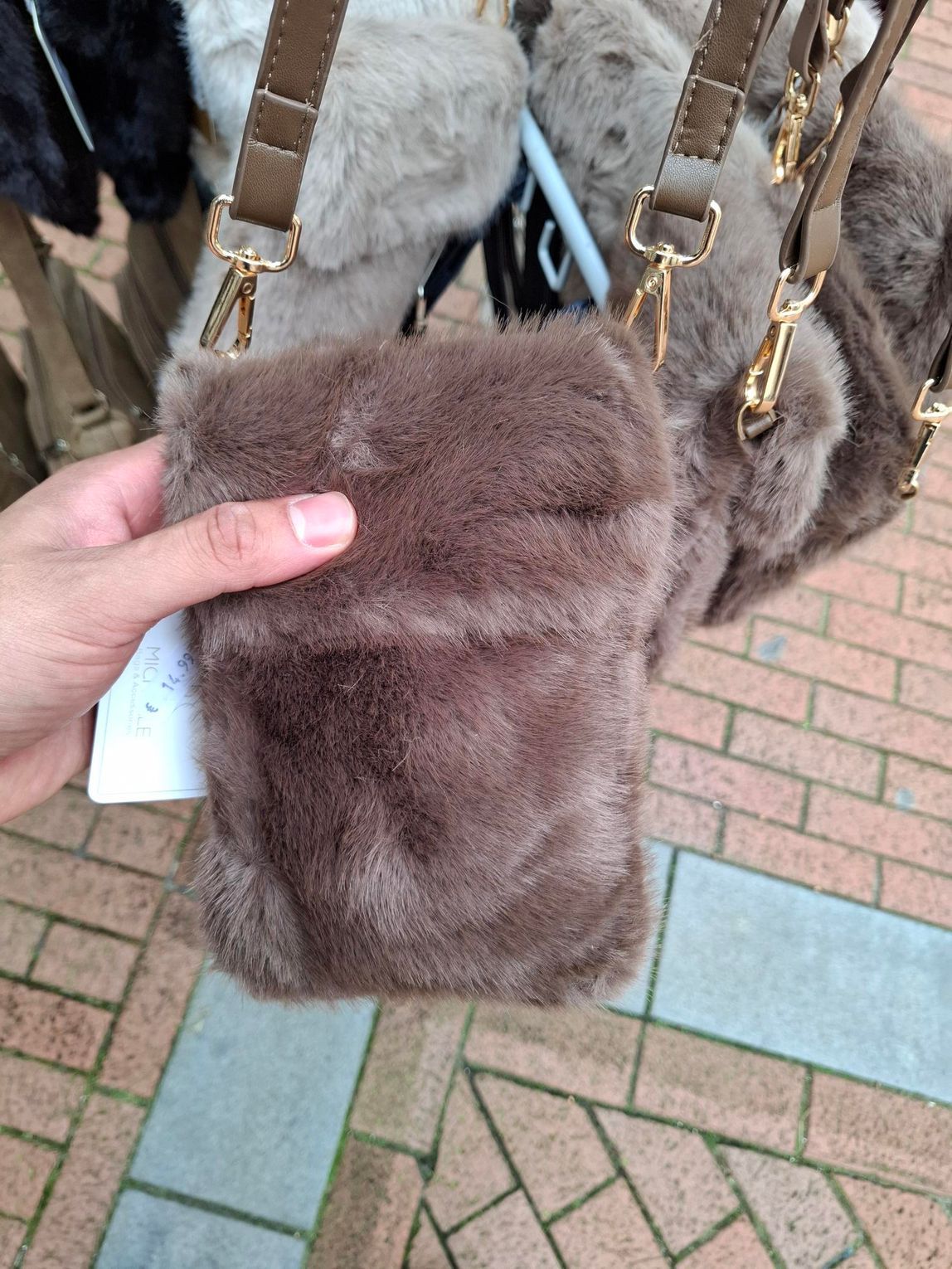 Hand die een bruine handtas van imitatiebont met goudkleurige details vasthoudt op een bakstenen stoep.