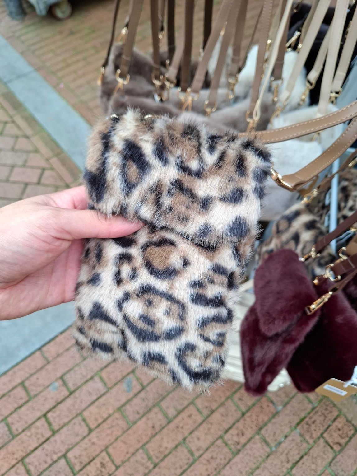 Hand die een accessoire van imitatiebont met luipaardprint en bordeauxrode kwastjes vasthoudt, zittend op een gevlochten stoel buiten.