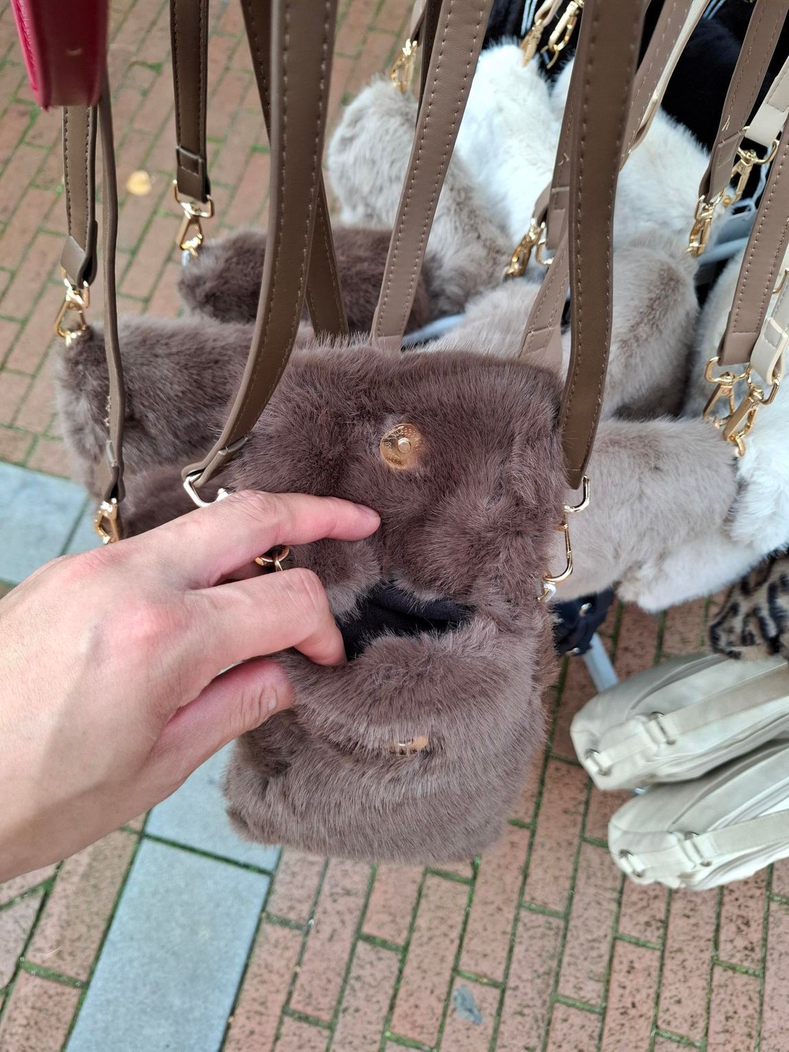 Hand die een grijze bonten tas met een schouderband vasthoudt op een stenen pad.