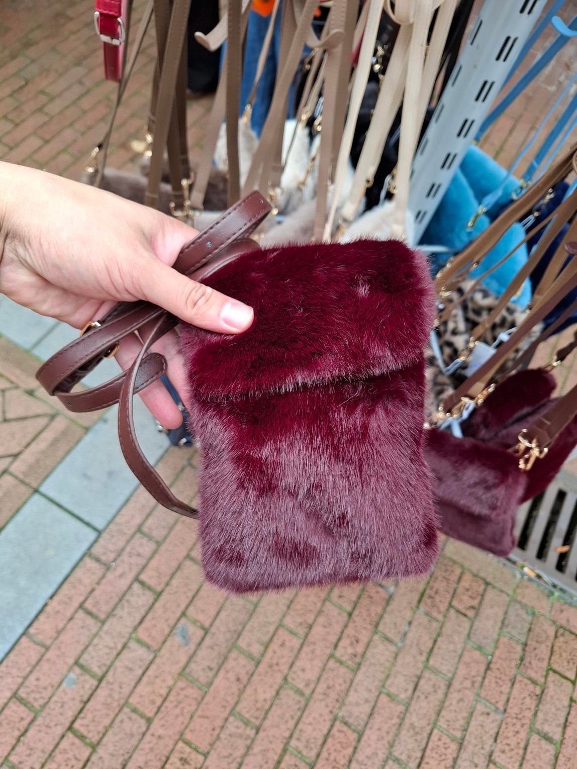 Hand met een bordeauxrode handtas van imitatiebont met schouderband op een stenen pad vlakbij een fietsenrek.