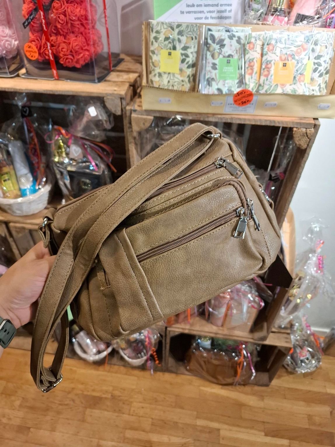Hand die een beige schoudertas vasthoudt in een cadeauwinkel, met kaarten en ingepakte artikelen erachter.