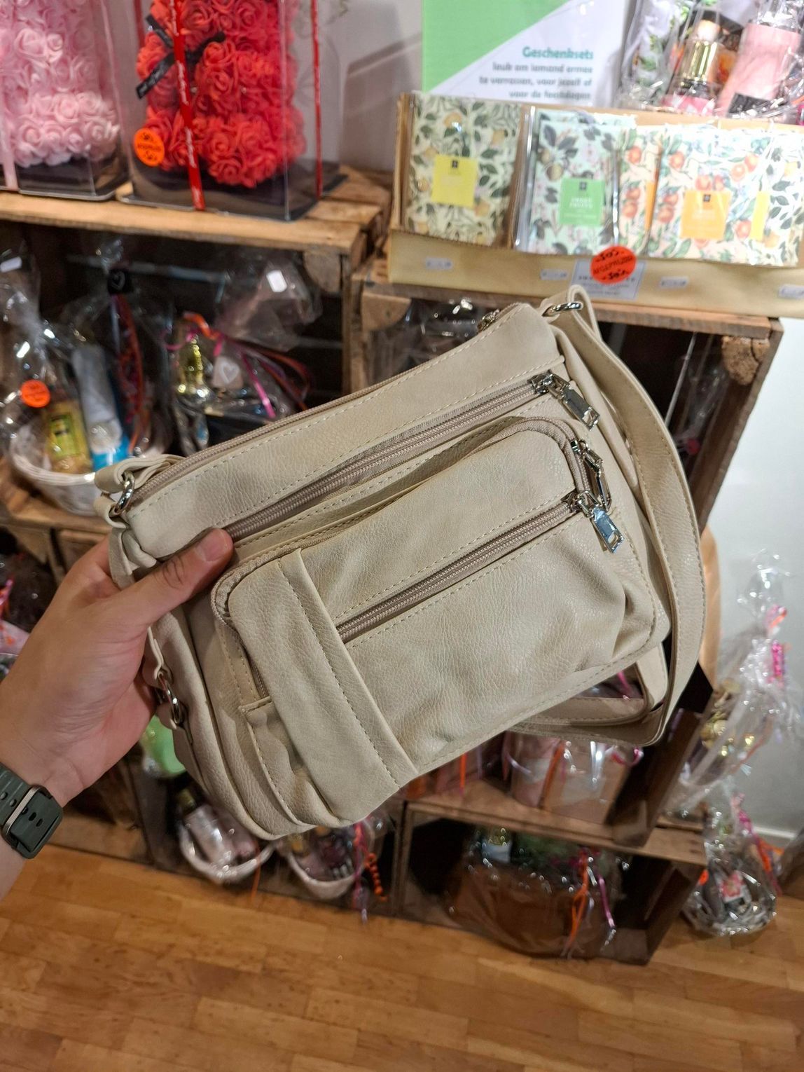 Hand die een beige canvas schoudertas vasthoudt in een winkel met schappen vol diverse artikelen.