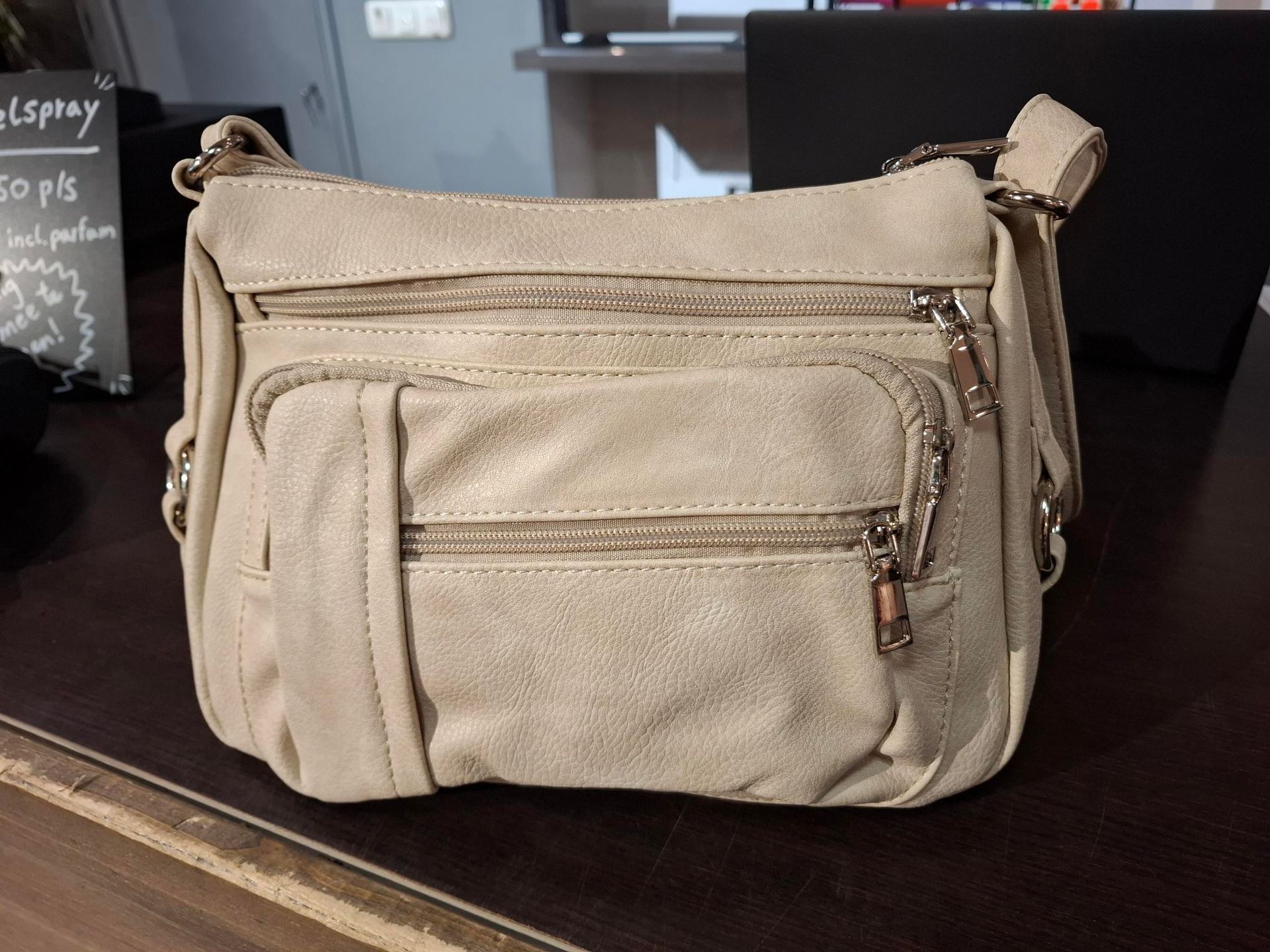 Hand die een beige canvas schoudertas vasthoudt in een winkel met schappen vol diverse artikelen.