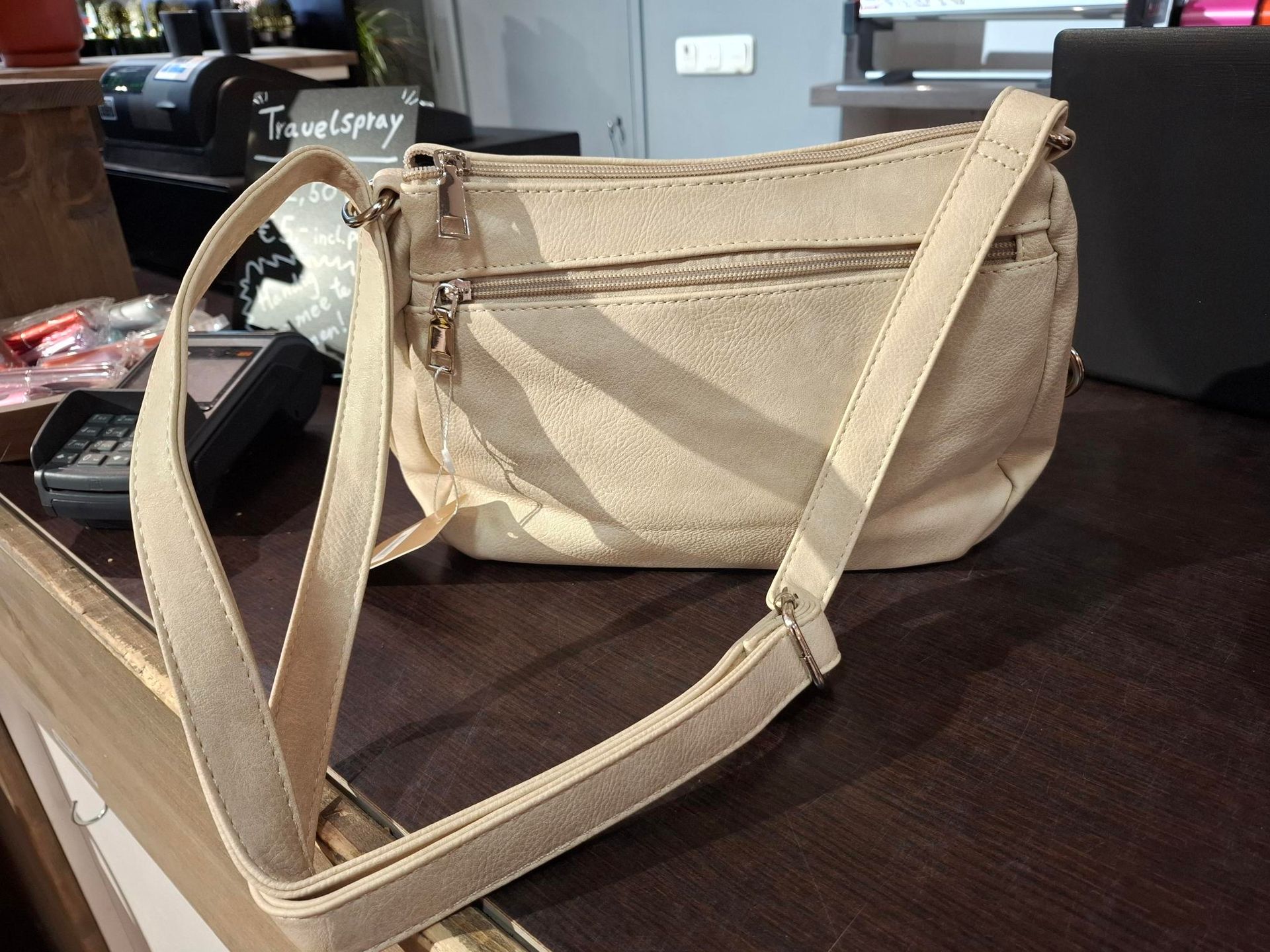 Hand die een beige schoudertas vasthoudt in een cadeauwinkel, met kaarten en ingepakte artikelen erachter.