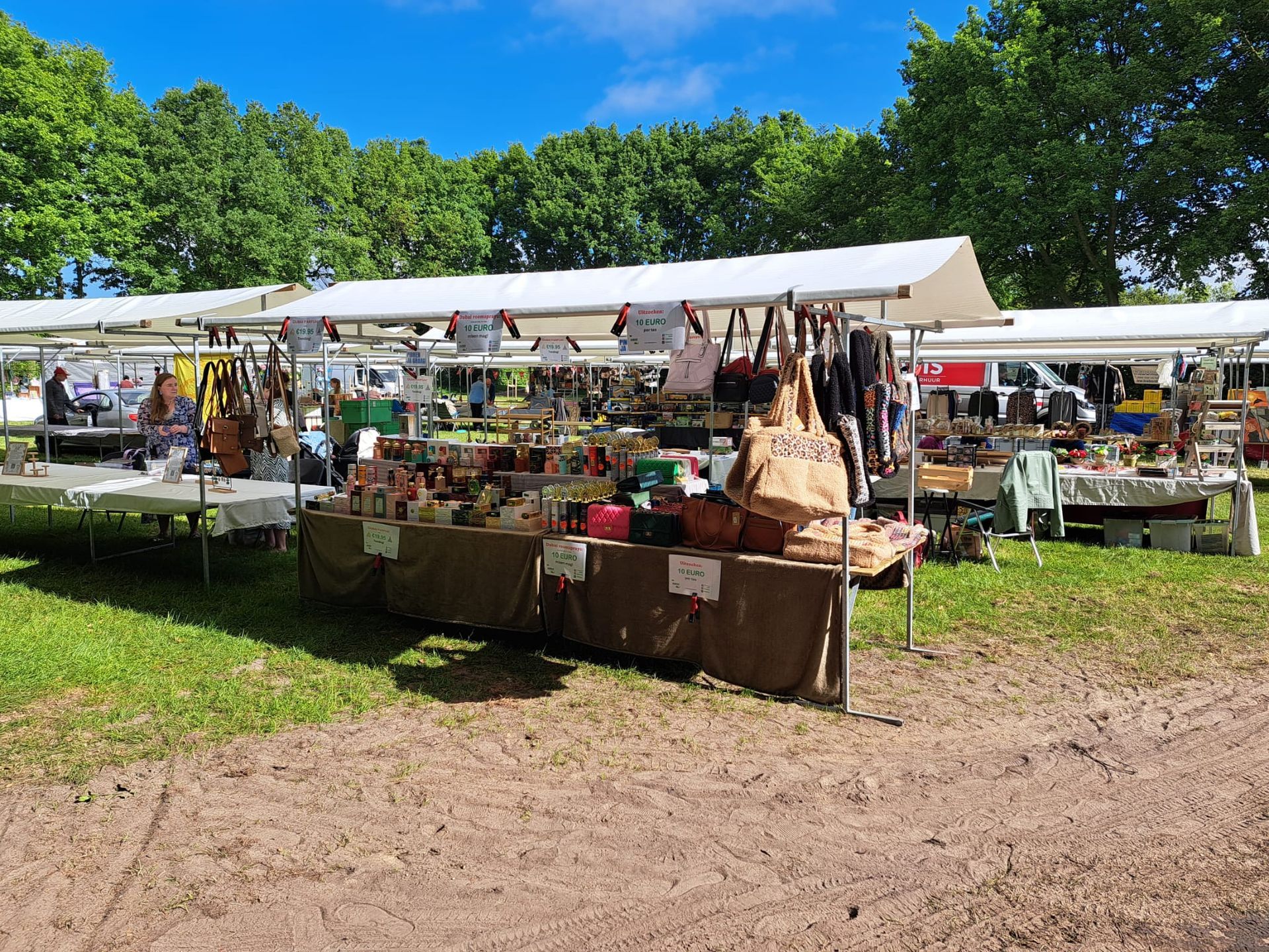 Op een marktkraam staan diverse tassen, accessoires en flessen uitgestald op een tafel onder een geel-wit gestreept afdak.