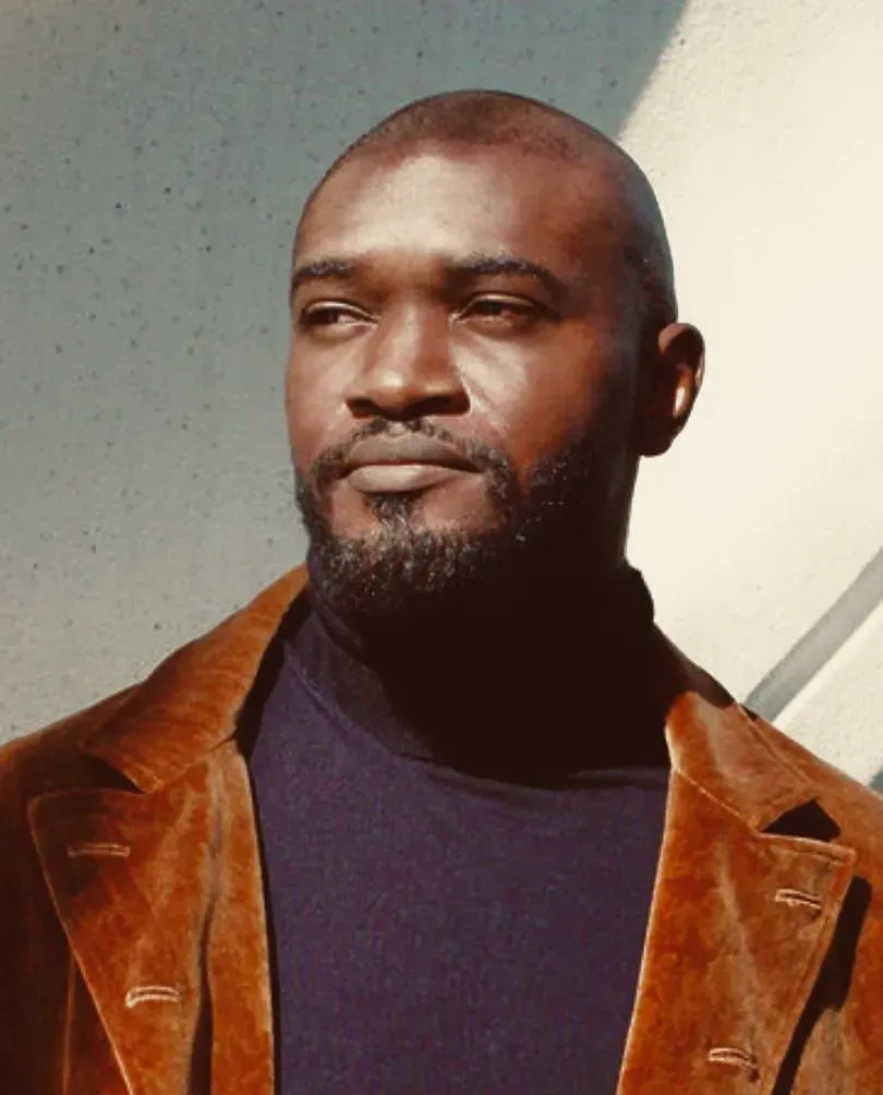 Man in brown jacket and black turtleneck, looking to the side, against a white wall.