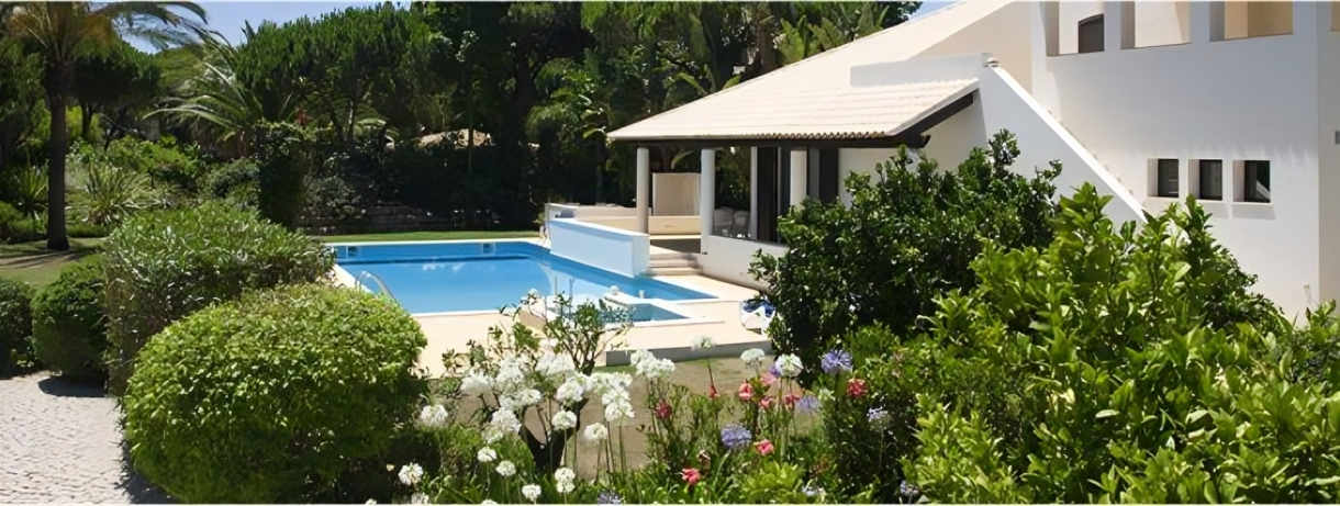 A white building with a pool surrounded by lush greenery under a bright sky.