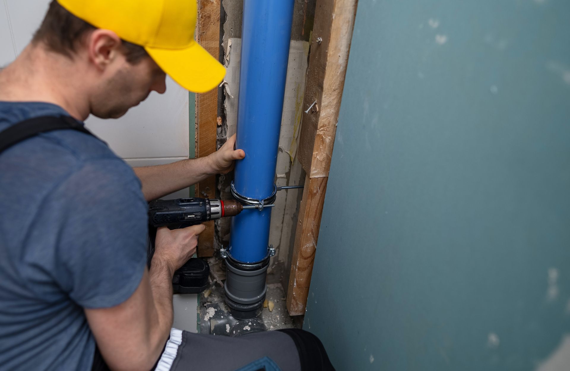 Man in yellow cap uses a drill to secure blue pipe against a wall.