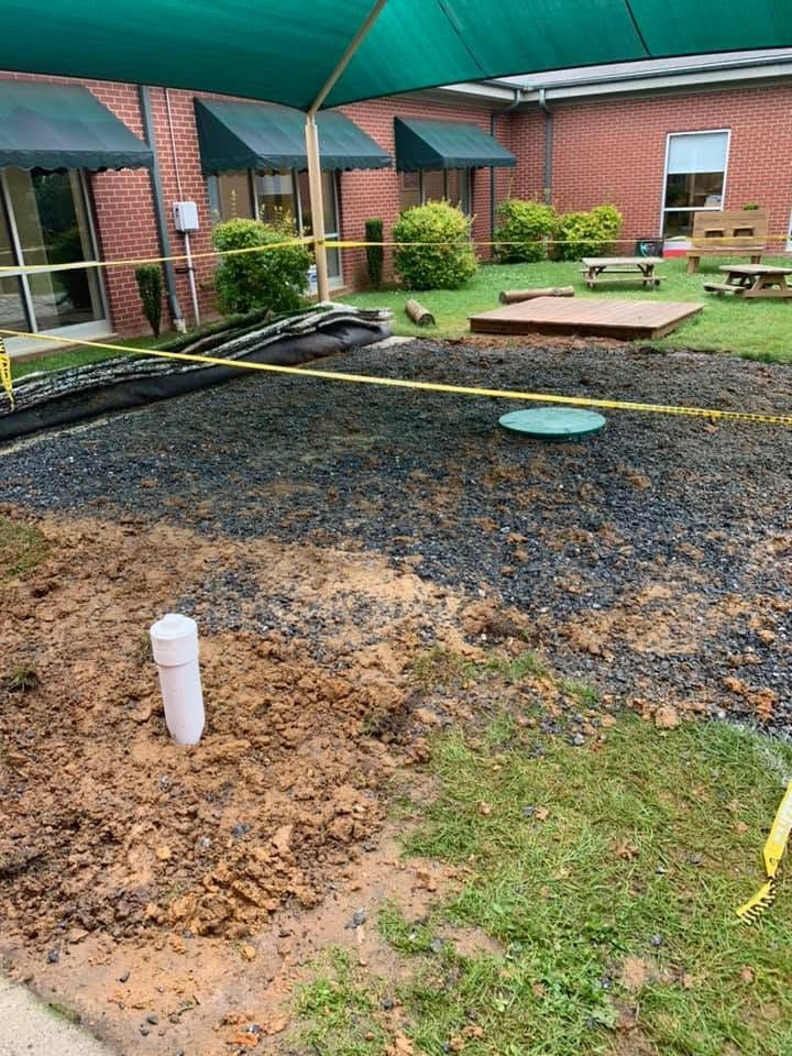 Partially dug-up lawn with a white pipe, gravel area, and brick building with green awnings in the background