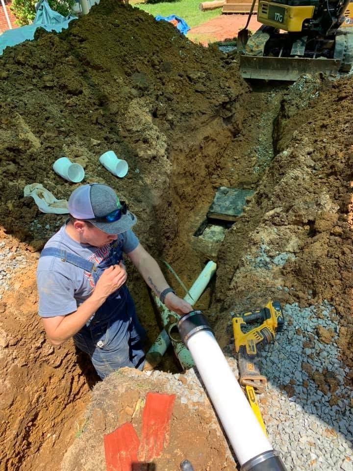 Worker in a trench connecting a large white pipe to underground utility lines.