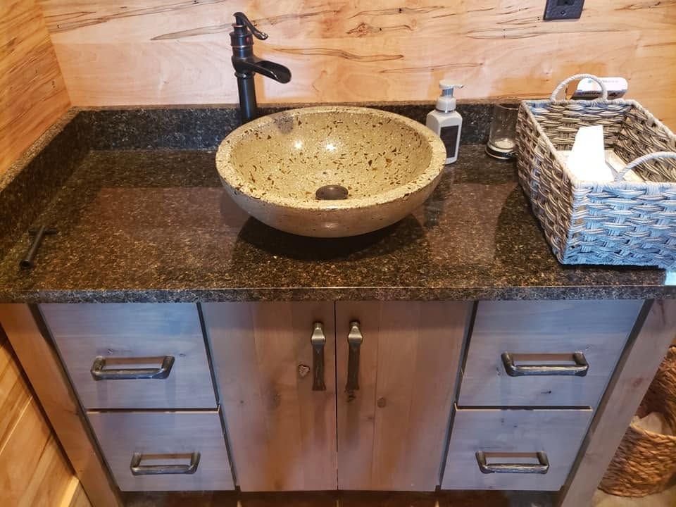 Bathroom vanity with dark granite countertop, stone vessel sink, and wicker tissue basket above wooden cabinets