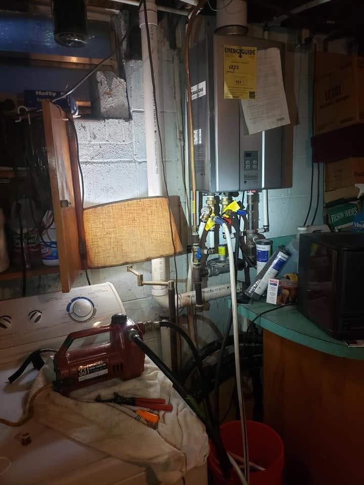 Cluttered basement utility area with pipes, electrical panel, broom, lamp, and cleaning supplies