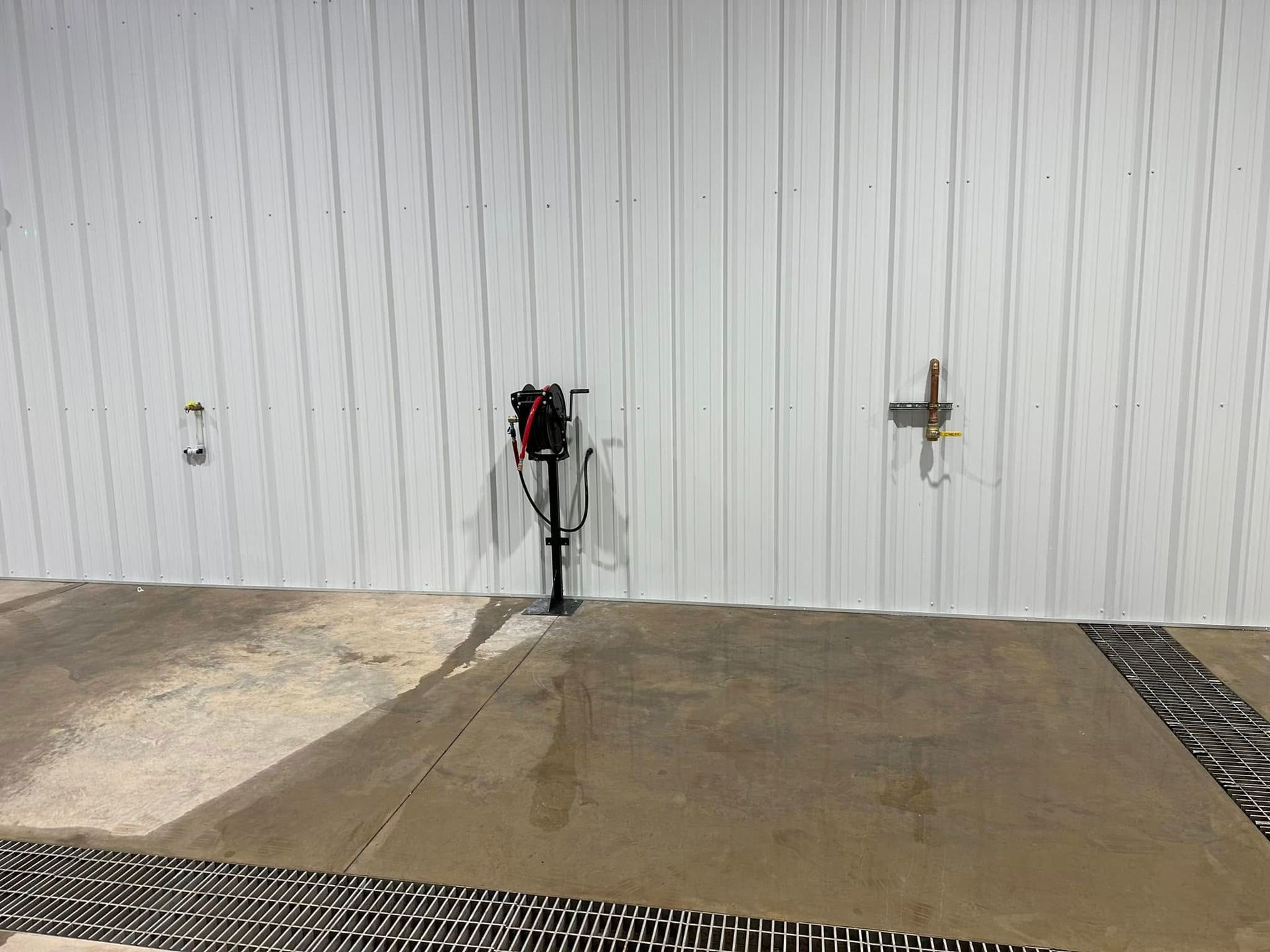 White metal wall with two hose bibs and a central hose reel above a concrete pad with drains.