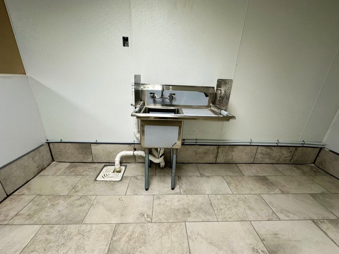 Empty room with a wall-mounted stainless steel sink and a small foot pedal on the floor.