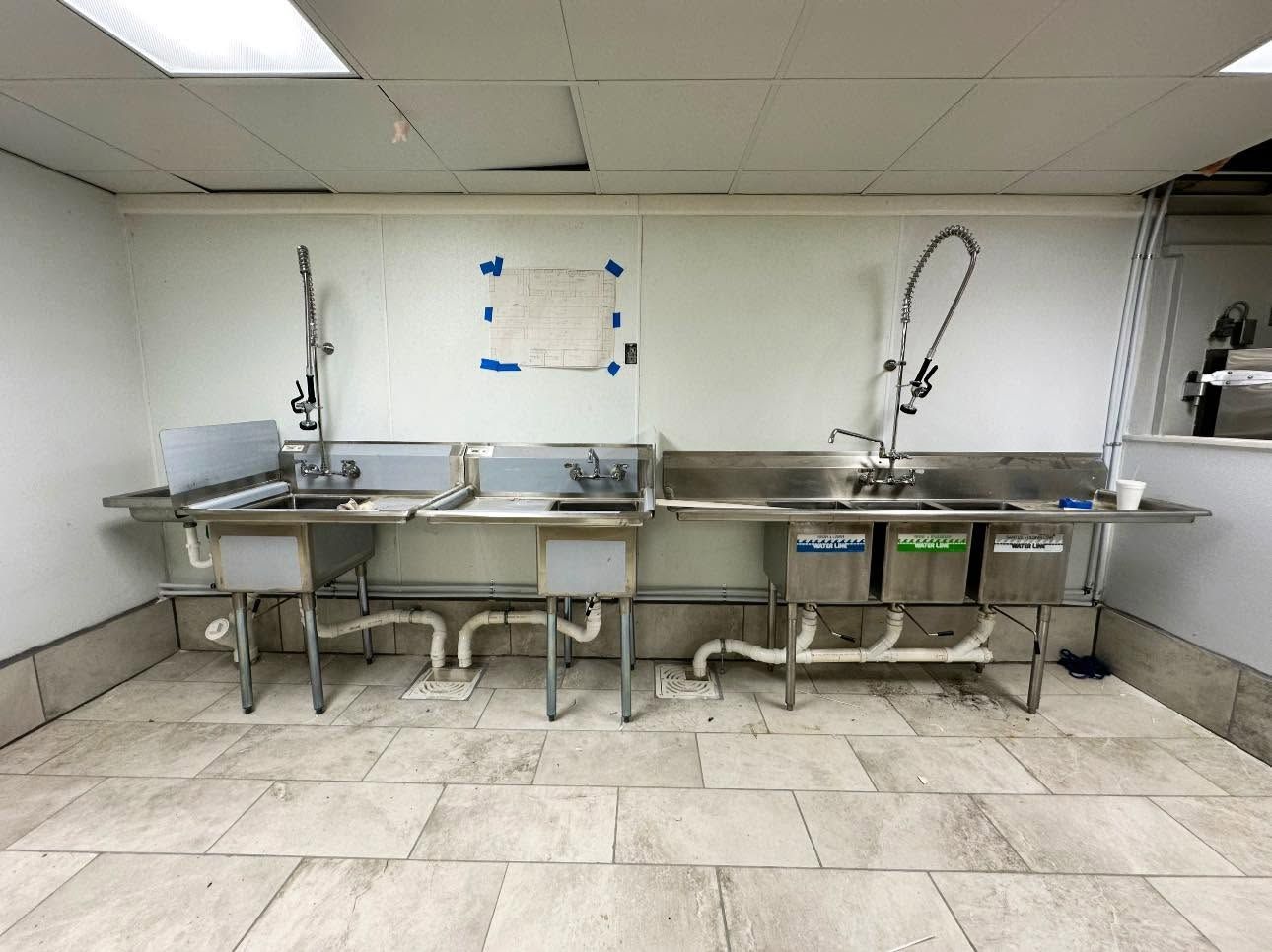 Commercial kitchen sink area with three stainless steel sinks and faucets along a tiled wall.