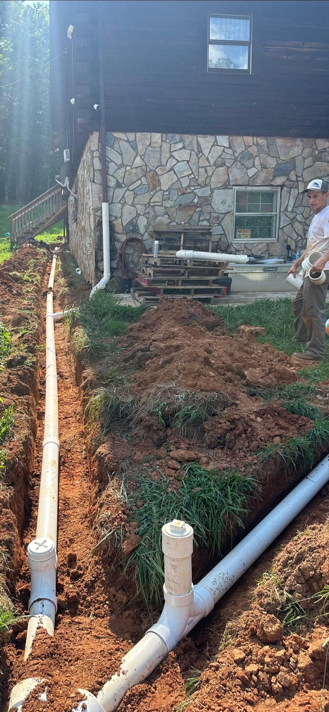 Steep muddy yard beside a stone house, with exposed pipes and drainage tubing downhill.