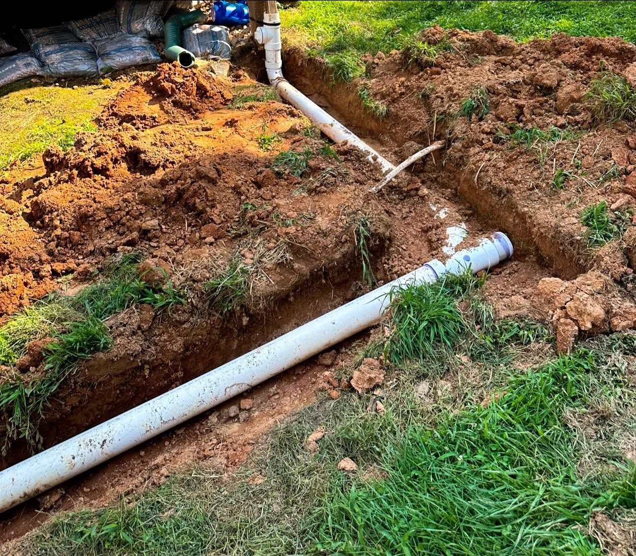 Broken white pipe leaking water in a muddy trench beside grass and soil.